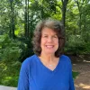 image of Juliet Haarbauer-Krupa wearing a blue shirt and the background is outside with a lot of trees.