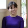 A woman with dark brown hair styled in a casual updo is smiling warmly. She is wearing a pearl necklace and a purple long-sleeved dress. She stands near a window with a stained-glass panel in the background, which features green and red accents. The lighting is soft and natural, casting a gentle glow on her face.