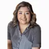 A woman with medium-length wavy brown hair with light highlights, wearing a short-sleeved gray button-up shirt. She is smiling and posed against a plain white background.