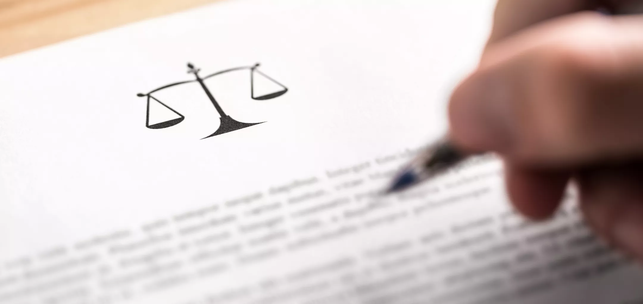 A close-up image of a person signing a legal document, with a prominent black symbol of balanced scales of justice printed at the top of the page. The text on the document is blurred, emphasizing the act of signing and the symbol representing law and fairness.
