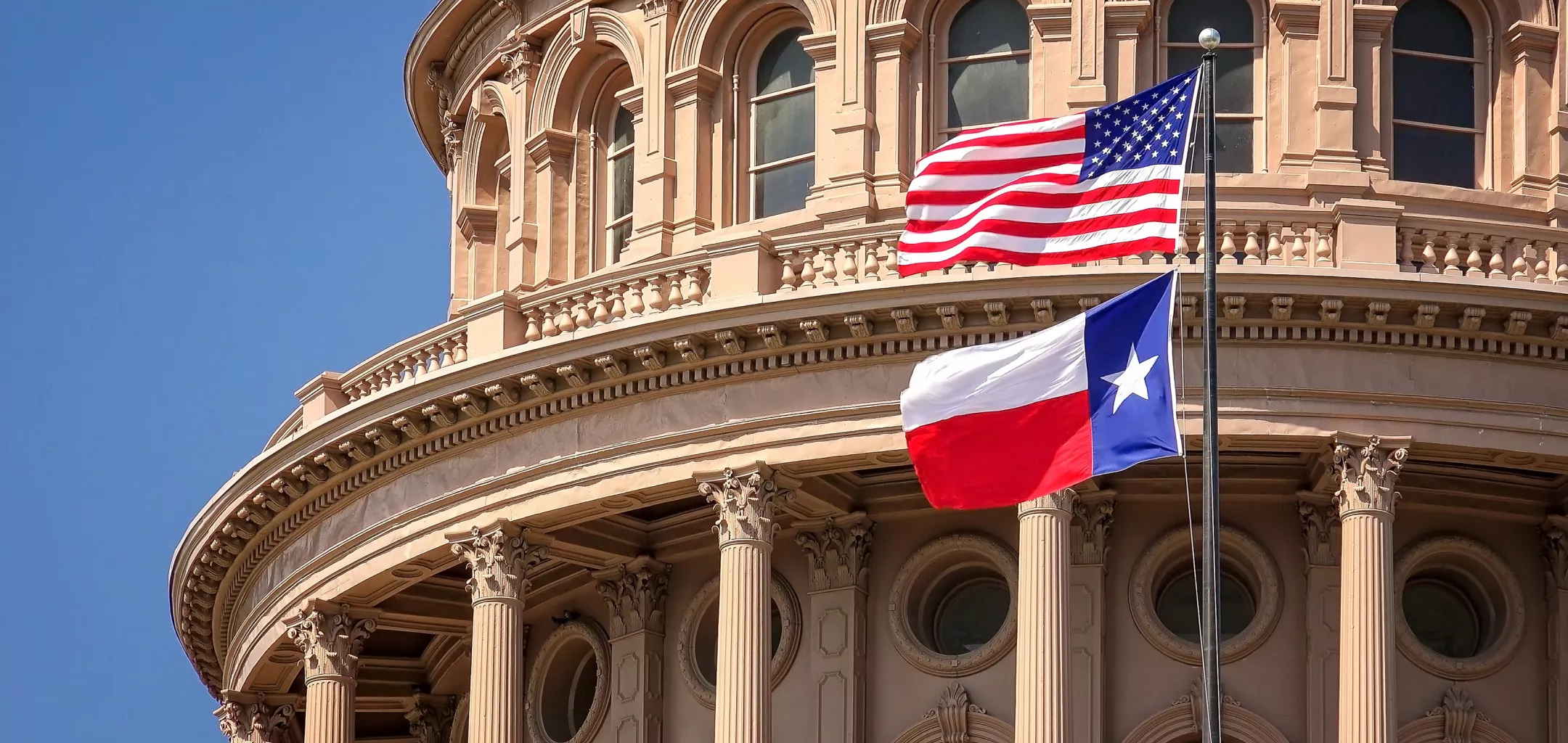 This image shows the upper portion of the Texas State Capitol building in Austin, Texas. Prominently displayed in front of the building are the United States flag and the Texas state flag, both flying on flagpoles. The architectural style includes classical columns and arched windows, characteristic of the capitol building’s historic design.