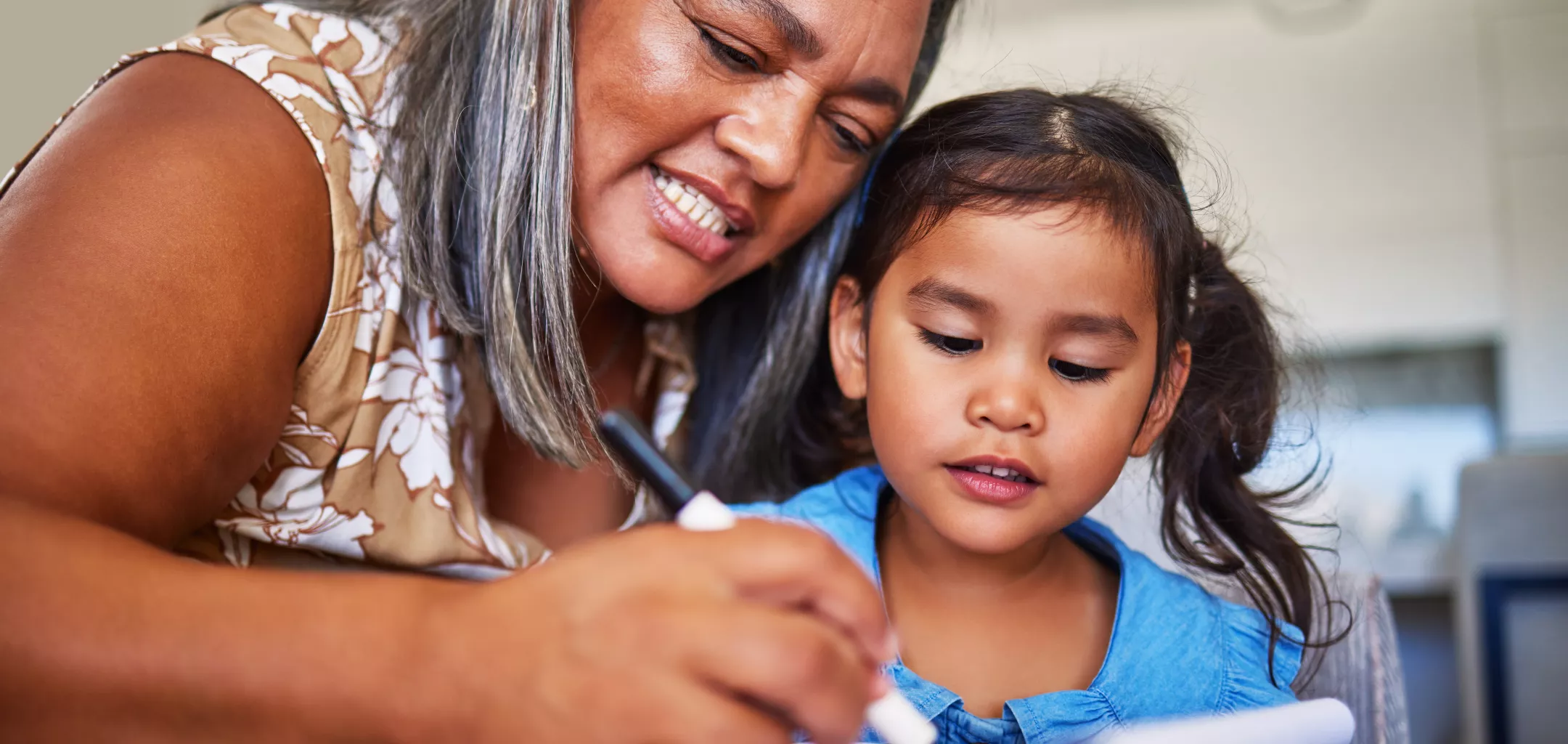 An adult woman with gray-streaked hair is sitting closely beside a young girl, guiding her hand as they draw or write with a marker on a notebook. The woman is smiling and focused, while the child looks intently at the paper. They appear to be engaged in a learning or creative activity at a table in a well-lit room.