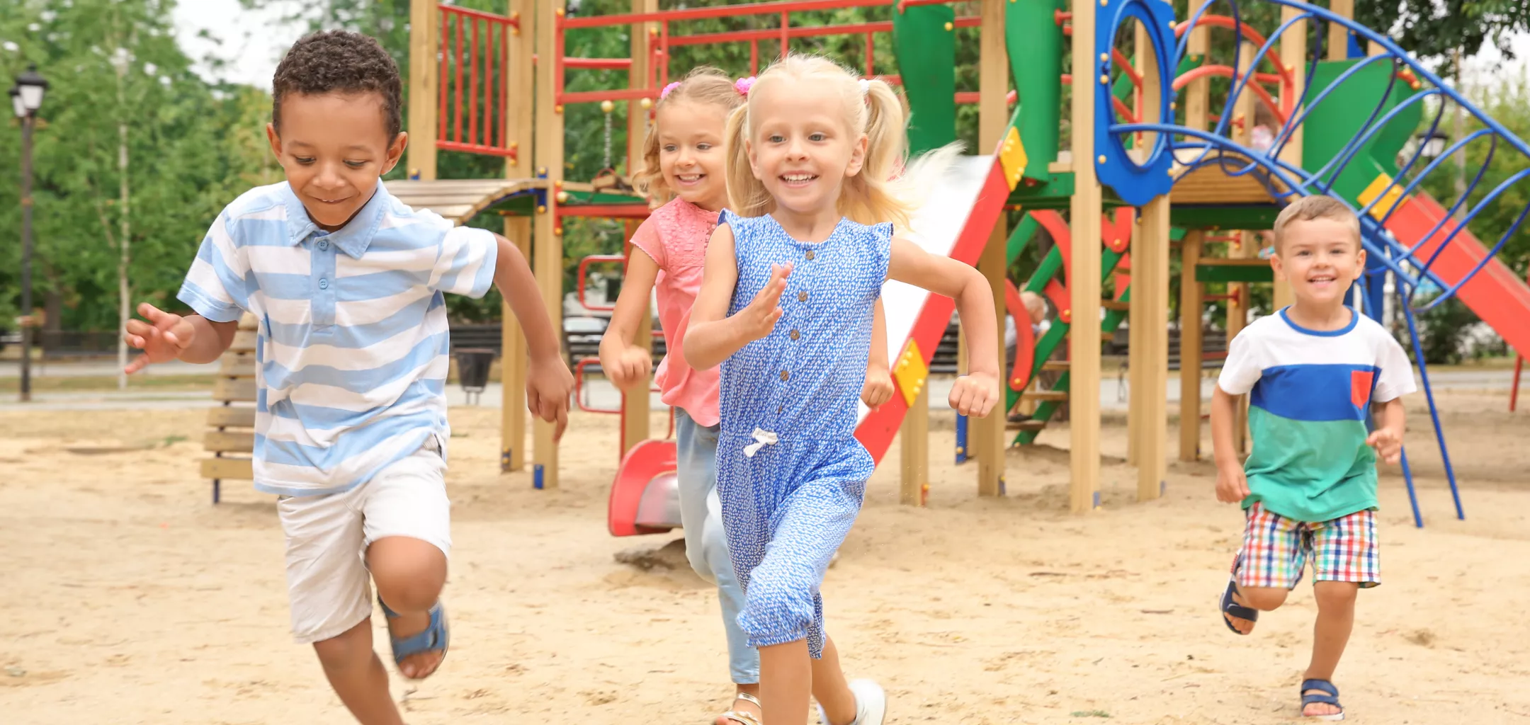 Four young children run happily on a sandy playground in front of colorful play structures, including slides and climbing equipment, on a sunny day.
